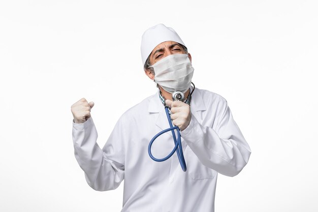 Vue de face de l'homme médecin en costume médical blanc portant un masque en raison du coronavirus tenant un stéthoscope et chantant sur un bureau blanc