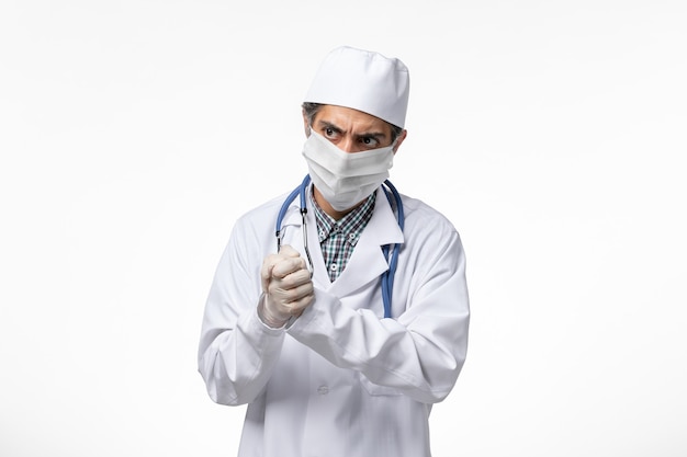 Vue de face de l'homme médecin en costume médical blanc en raison d'un coronavirus portant un masque sur une surface blanche
