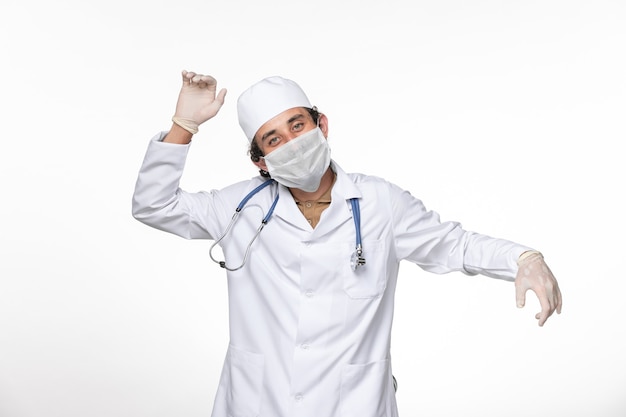 Photo gratuite vue de face de l'homme médecin en costume médical avec masque comme protection contre le covid- légèrement danser sur la pandémie de coronavirus virus splash mur blanc