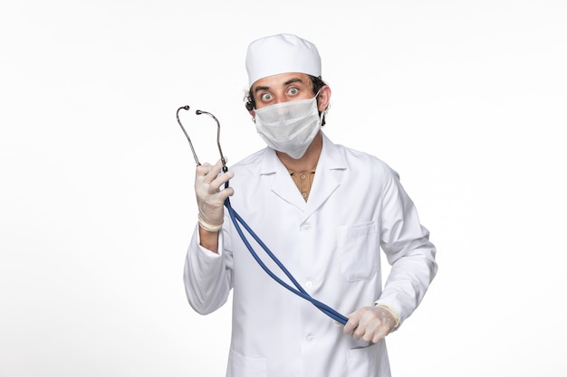 Vue de face de l'homme médecin en costume médical avec masque comme protection contre le stéthoscope covid- holding sur la pandémie de coronavirus splash de virus de bureau blanc