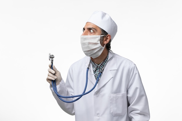 Vue de face de l'homme médecin en costume médical et masque en raison de coronavirus tenant un stéthoscope sur un bureau blanc