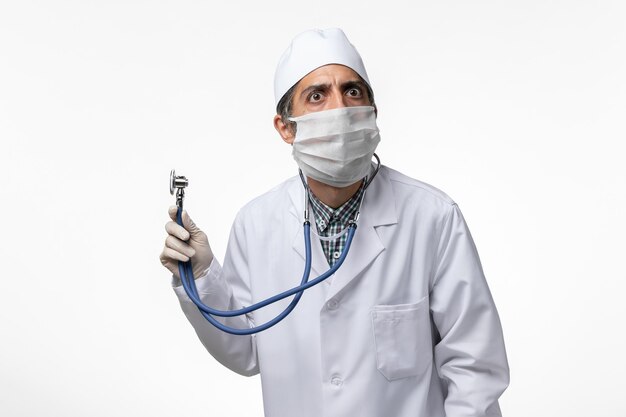 Vue de face de l'homme médecin en costume médical et masque en raison du coronavirus tenant un stéthoscope sur une surface blanche