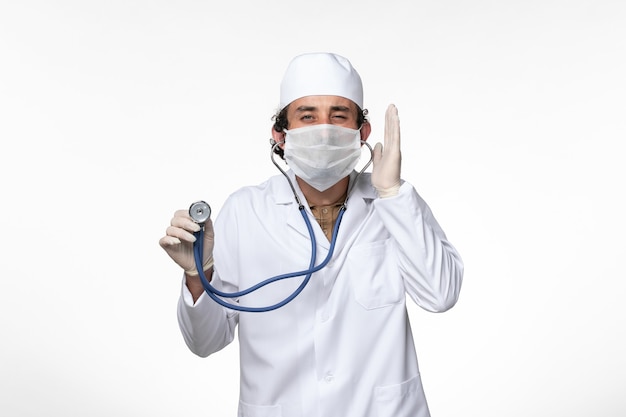 Vue de face de l'homme médecin en costume médical et portant un masque comme une protection contre les covid- à l'aide d'un stéthoscope sur la pandémie de covid maladie virus mur blanc