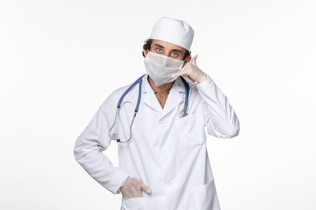 Vue de face de l'homme médecin en costume médical portant un masque stérile comme protection contre le covid montrant un appel téléphonique pose sur un bureau blanc maladie pandémique du coronavirus