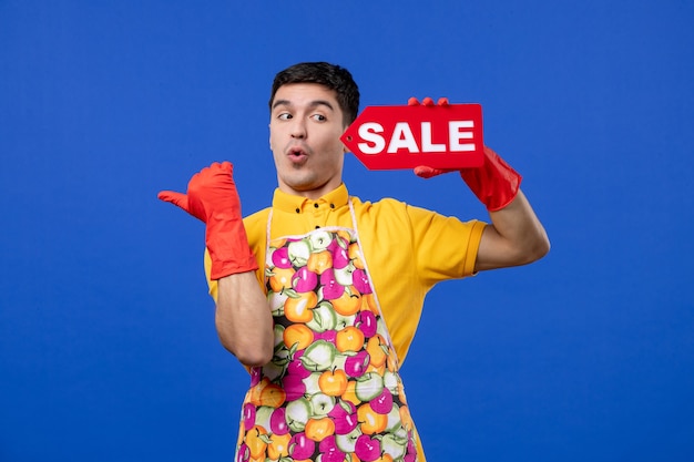 Vue de face de l'homme de ménage demandé avec des gants de vidange rouges tenant une pancarte de vente pointant vers la gauche sur le mur bleu