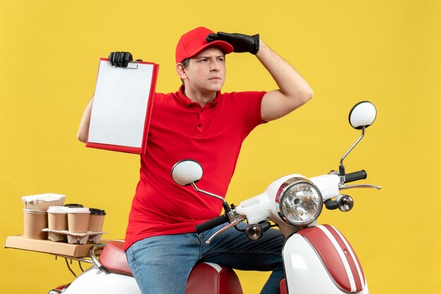 Vue de face de l'homme de messagerie ciblé portant chemisier rouge et gants de chapeau en masque médical délivrant la commande assis sur le document tenant le scooter