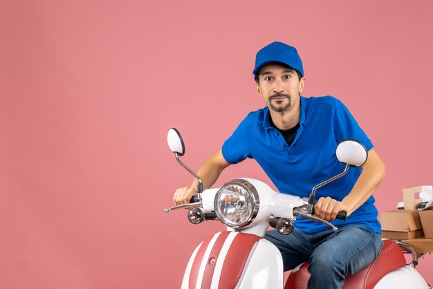 Vue de face d'un homme de messagerie heureux portant un chapeau assis sur un scooter sur fond pêche pastel