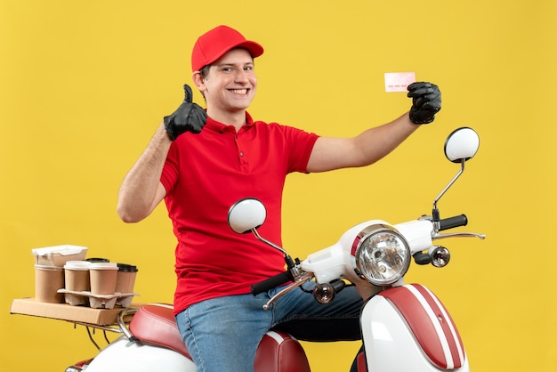 Vue de face de l'homme de messagerie portant chemisier rouge et gants de chapeau dans un masque médical délivrant l'ordre assis sur un scooter tenant une carte bancaire faisant un geste ok