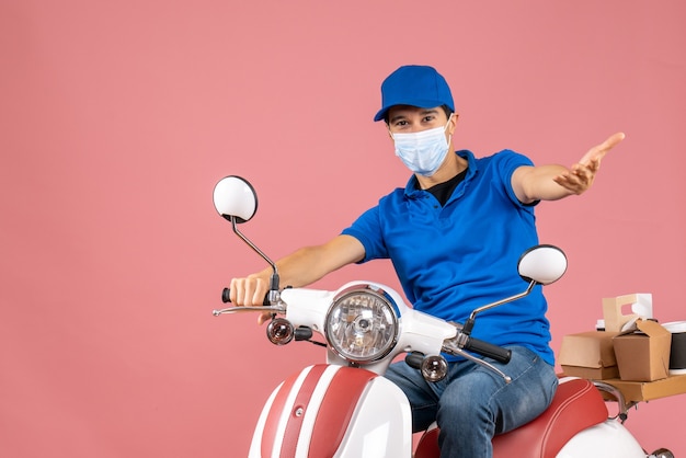 Vue de face de l'homme de messagerie se demandant dans un masque médical portant un chapeau assis sur un scooter livrant des commandes sur fond de pêche pastel