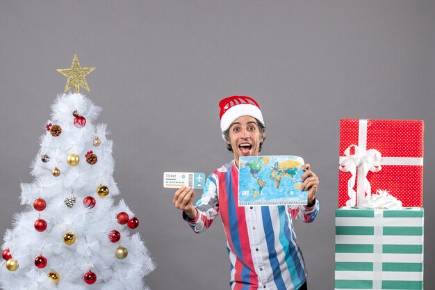 Vue de face homme de Noël heureux avec bonnet de noel printemps en spirale tenant carte du monde et billet de voyage