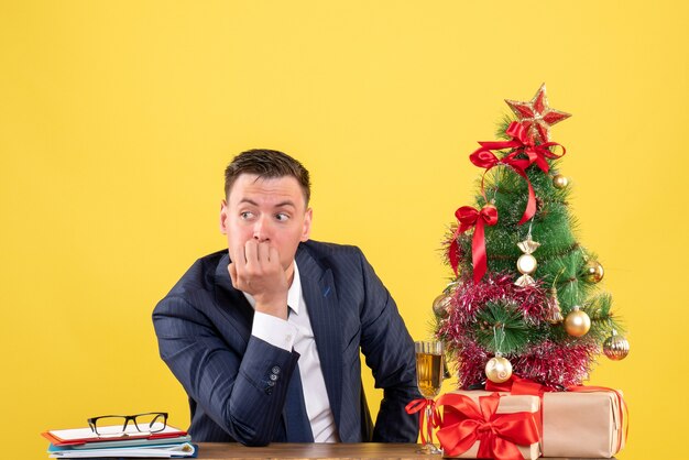 Vue de face de l'homme réfléchi assis à la table près de l'arbre de Noël et présente sur jaune