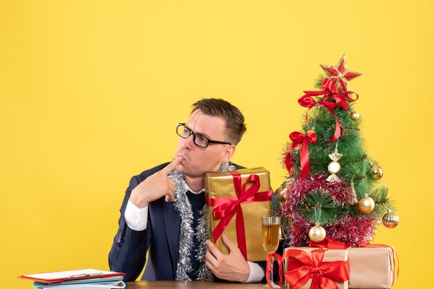 Vue de face de l'homme réfléchi mettant le doigt sur la bouche assis à la table près de l'arbre de Noël et présente sur jaune