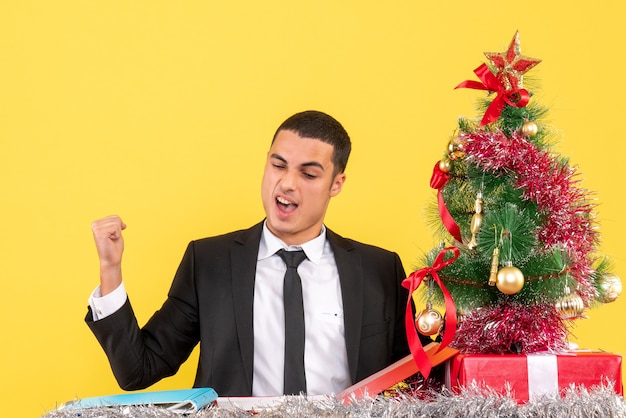 Vue de face homme satisfait assis à la table montrant l'arbre de Noël de geste gagnant et cadeaux