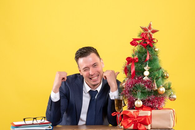 Vue de face de l'homme souriant pointant sur l'arbre de Noël assis à la table près de l'arbre de Noël et cadeaux sur jaune