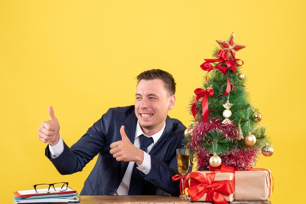 Vue de face de l'homme sourit faisant le pouce vers le haut signe assis à la table près de l'arbre de Noël et des cadeaux sur jaune