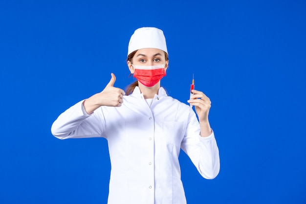 Vue de face infirmière en costume médical blanc avec masque rouge et injection dans ses mains sur bleu