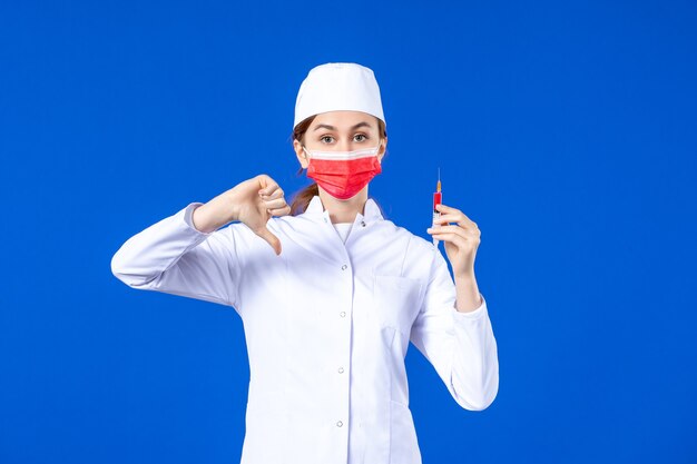 Vue de face infirmière en costume médical blanc avec masque rouge et injection dans ses mains sur bleu