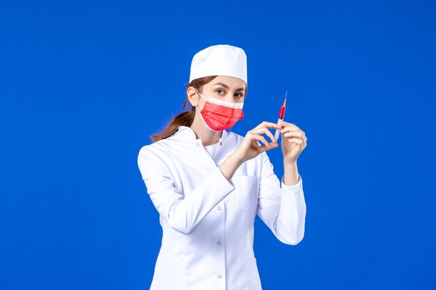 Vue de face infirmière en costume médical blanc avec masque rouge et injection dans ses mains sur bleu