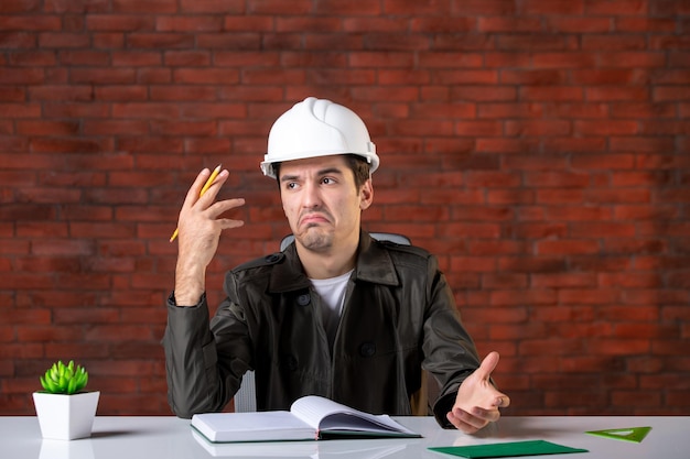 Vue de face ingénieur masculin assis derrière son lieu de travail dans un casque blanc document entreprise propriété d'entreprise plan de l'entrepreneur constructeur d'emplois