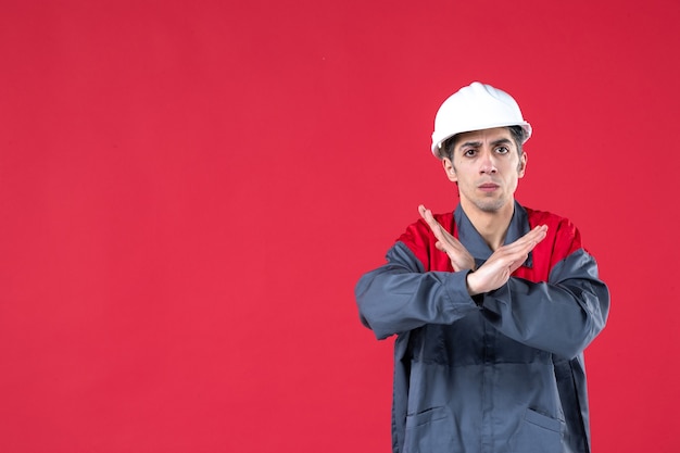 Vue de face d'un jeune architecte nerveux en uniforme avec un casque et faisant un geste d'arrêt sur un mur rouge isolé