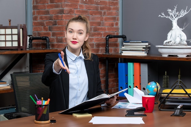 Photo gratuite vue de face d'une jeune assistante confiante assise à son bureau et tenant un document accueillant quelqu'un au bureau