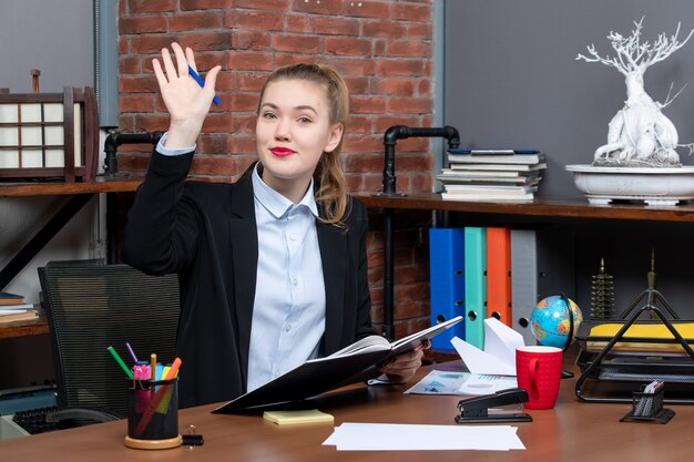 Vue de face d'une jeune assistante confiante assise à son bureau et tenant un document disant bonjour à quelqu'un au bureau