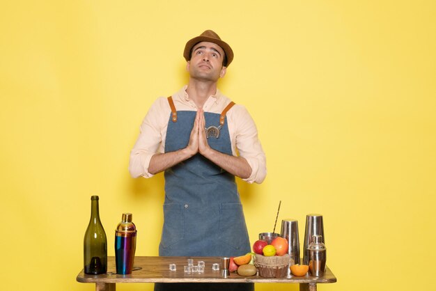 Vue de face jeune barman masculin devant la table avec des boissons shakers sur fond jaune