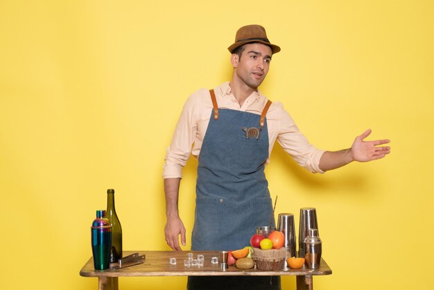 Vue de face jeune barman masculin devant la table avec des boissons shakers sur fond jaune