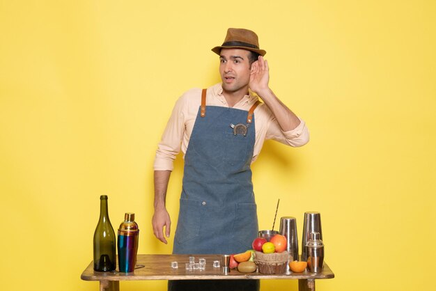 Vue de face jeune barman masculin devant la table avec des boissons shakers sur fond jaune