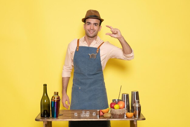 Vue de face jeune barman masculin devant la table avec des boissons shakers sur fond jaune