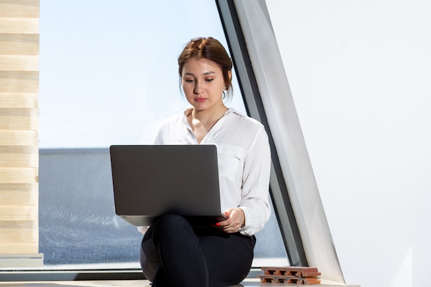 Une Vue De Face Jeune Belle Dame En Chemise Blanche Pantalon Noir Assis Près De La Fenêtre Travaillant Sur L'ordinateur Portable Pendant La Journée