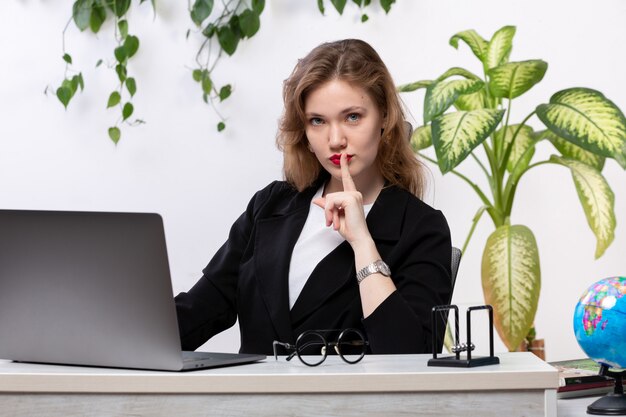 Une vue de face jeune belle dame en chemise blanche et veste noire à l'aide de son ordinateur portable en face de table souriant montrant signe de silence avec des feuilles suspendues