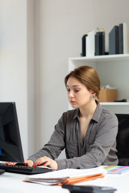 Une vue de face jeune belle dame en chemise grise travaillant avec les documents à l'aide de son pc assis à l'intérieur de son bureau pendant la journée