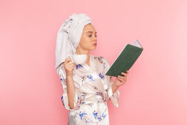 Une vue de face jeune belle dame en peignoir, boire du café et lire un livre