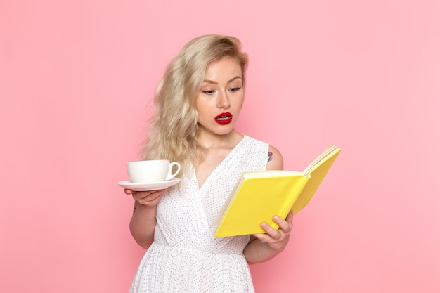 Photo gratuite une vue de face jeune belle dame en robe blanche de boire un thé en lisant un cahier