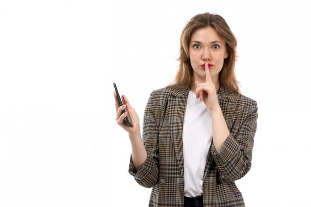 Une vue de face jeune belle dame en t-shirt blanc jeans noirs et manteau à l'aide de smartphone montrant signe de silence sur le blanc