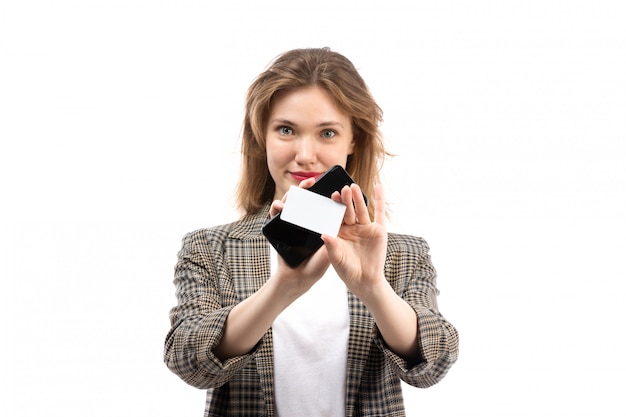 Une vue de face jeune belle dame en t-shirt blanc jeans noirs et manteau tenant un smartphone noir et une carte blanche souriant sur le blanc