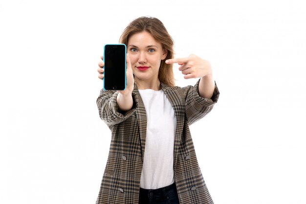 Une vue de face jeune belle dame en t-shirt blanc jeans noirs et manteau tenant smartphone souriant sur le blanc