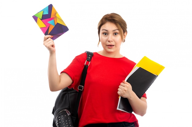 Une vue de face jeune belle dame en t-shirt rouge jeans noirs tenant différents cahiers et fichiers avec sac sur le blanc