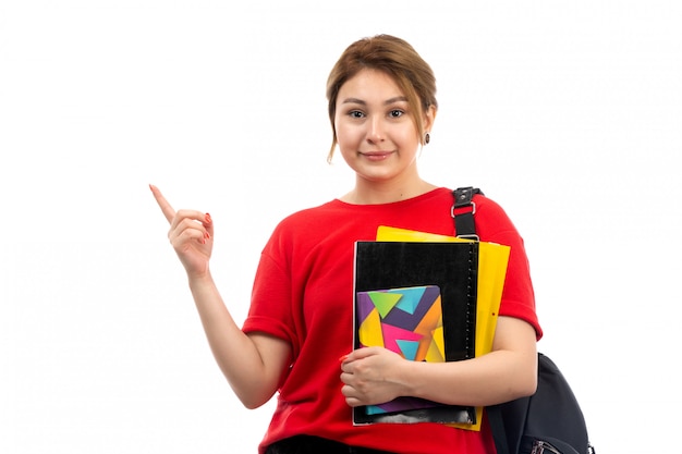 Une vue de face jeune belle dame en t-shirt rouge jeans noirs tenant différents cahiers et fichiers souriant avec sac sur le blanc