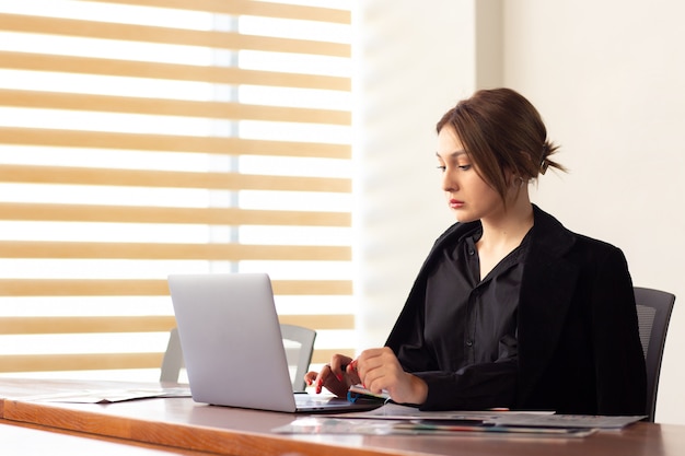 Une vue de face jeune belle femme d'affaires en chemise noire veste noire à l'aide de son ordinateur portable argent écrit lecture travaillant à l'intérieur de son travail de bureau bâtiment de travail