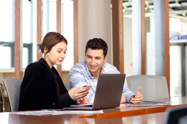 Une vue de face jeune belle femme d'affaires en chemise noire veste noire avec jeune homme regardant par téléphone en regardant quelque chose à l'intérieur de son travail de bureau bâtiment de l'emploi