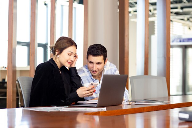 Une vue de face jeune belle femme d'affaires en chemise noire veste noire avec jeune homme vérifiant le téléphone en regardant quelque chose à l'intérieur de son travail de bureau