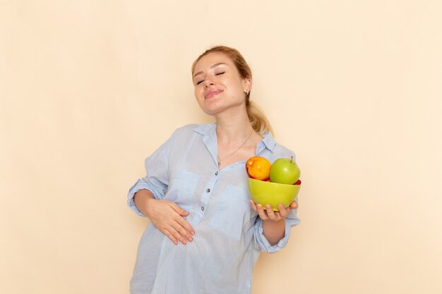 Vue de face jeune belle femme en chemise tenant la plaque avec des fruits et souriant posant sur le mur crème fruits modèle femme pose dame