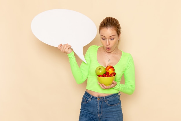 Photo gratuite vue de face jeune belle femme en chemise verte tenant une assiette pleine de fruits et signe blanc sur mur crème fruits modèle femme alimentaire vitamine