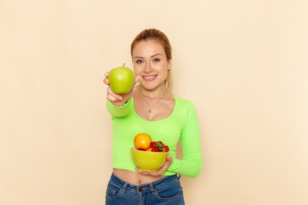 Vue de face jeune belle femme en chemise verte tenant la plaque avec des fruits et tenant la pomme verte sur le mur crème légère modèle de fruits femme pose