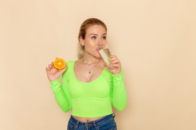 Vue de face jeune belle femme en chemise verte tenant une tranche d'orange et un verre d'eau potable sur le mur crème fruit modèle femme moelleux