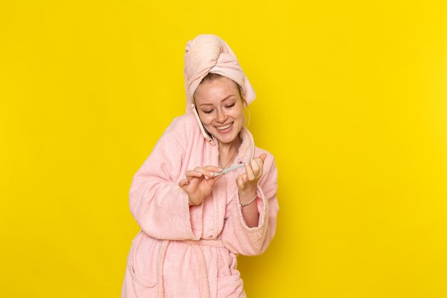 Une vue de face jeune belle femme en peignoir rose parlant au téléphone fixant ses ongles