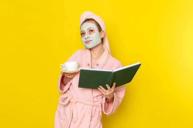 Une vue de face jeune belle femme en peignoir rose tenant livre vert et tasse de thé