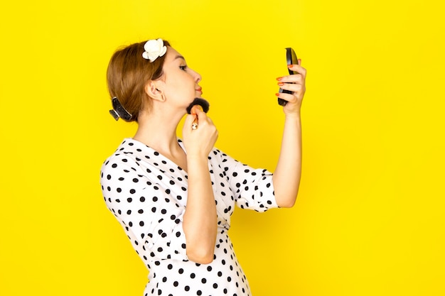 Une vue de face jeune belle femme en robe à pois noir et blanc faisant du maquillage sur jaune